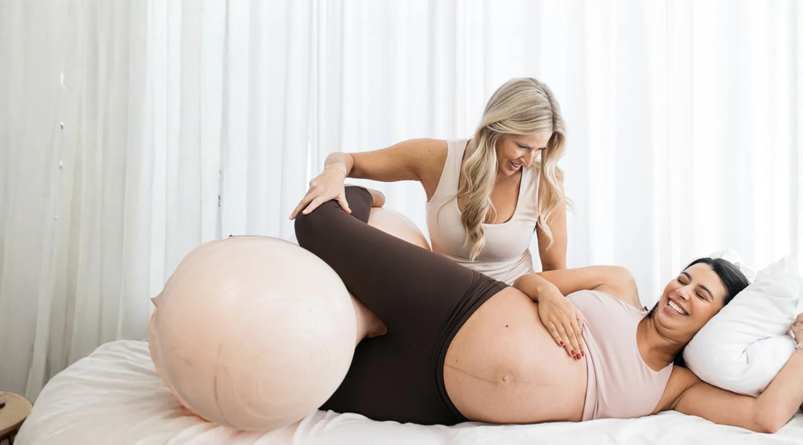 A Revelle childbirth educator coaches a smiling pregnant woman through a labor position with a peach birth ball, on a calm bright bed framed by soft white curtains